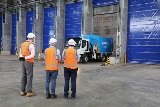 Three people wearing high visibility and safety helmets in warehouse with blue waste truck.