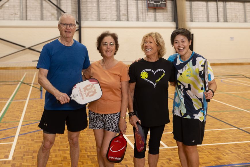 4 people stading together smiling with rackets at a pickleball court