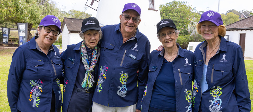 Group of five volunteers wearing hats and smiling in a park with an old mill in the background.