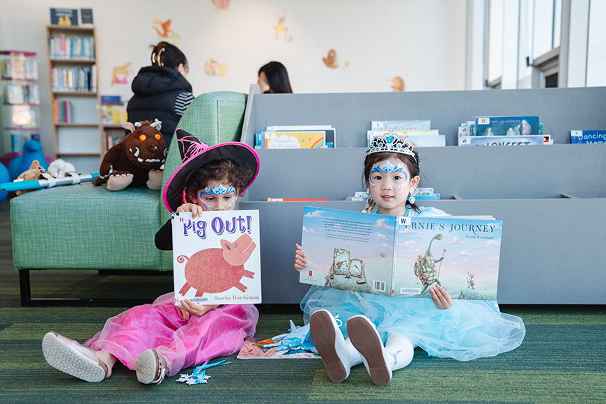 Two little girls in dress up sitting on the floor reading picture books.