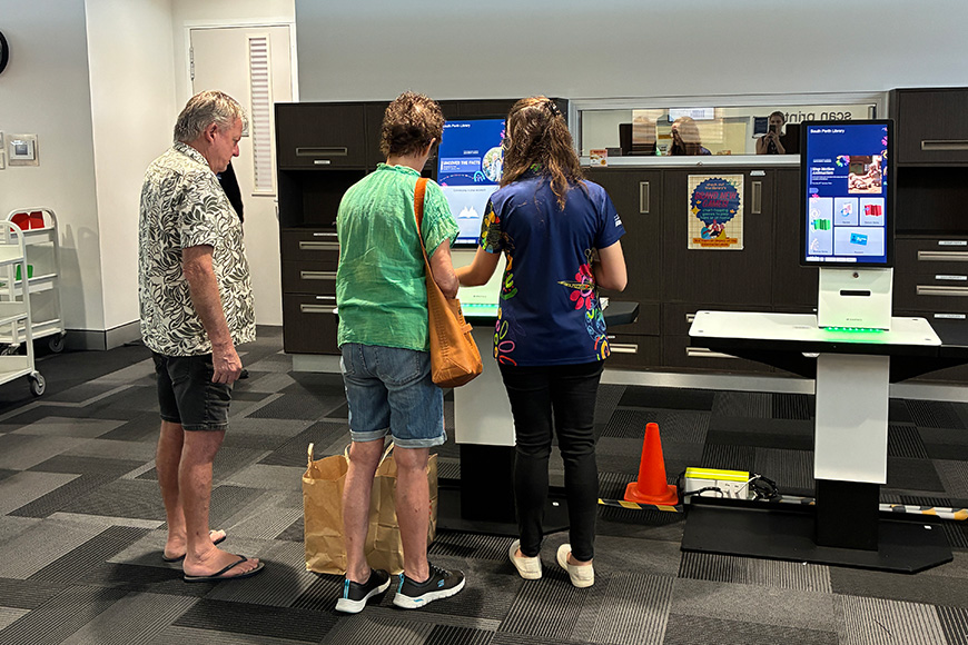 Three people standing in  front of self-service library kiosks