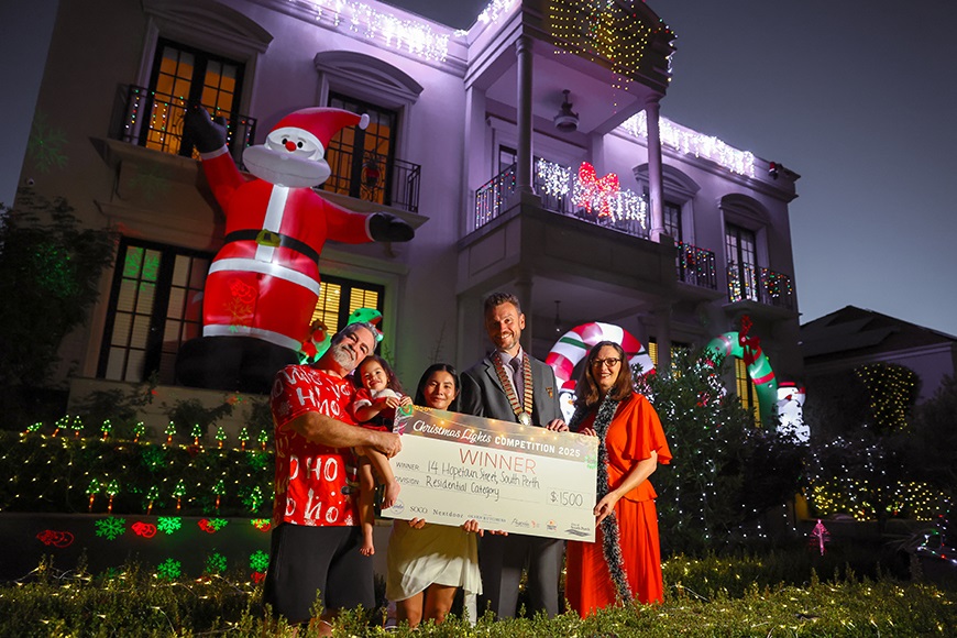 People smiling and holding a novelty cheque in front of a house covered in Christmas Lights