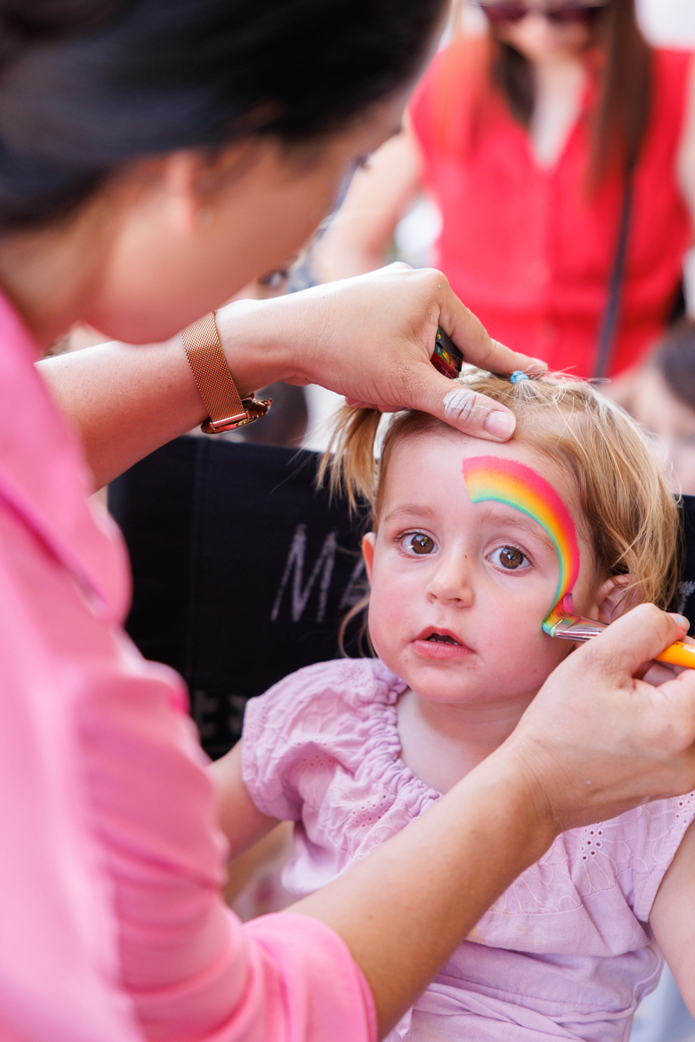 A young child getting a rainbow painted on their face.