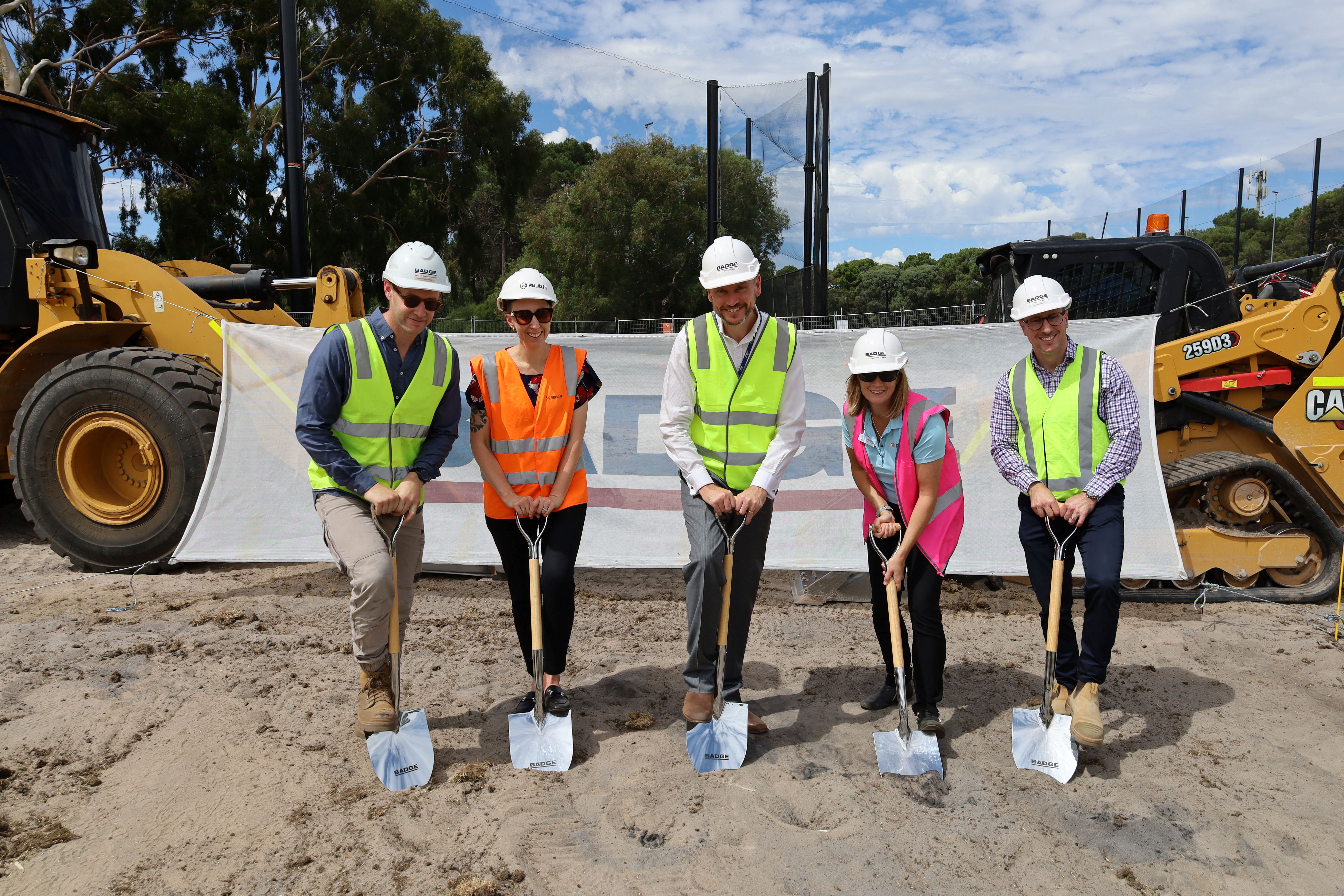 Mayor Greg Milner with two women and two men holding shovels and wearing hard hats and fluro vests in front of a construction truck