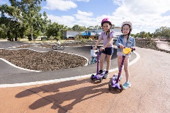 Two girls on scooters smiling at the camera with the bike track in the background