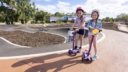 Twogirlsonscooterssmilingatthecamerawiththebiketrackinthebackground