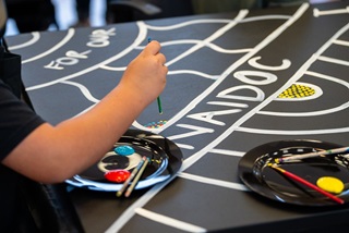 ChildhandholdingapaintbrushpaintingfirstnationsdotartworksurroundingthewordNAIDOC