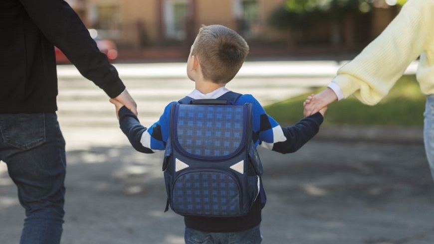 Ayoungchildwithaschoolbackpackholdinghandswithoneparenteachside