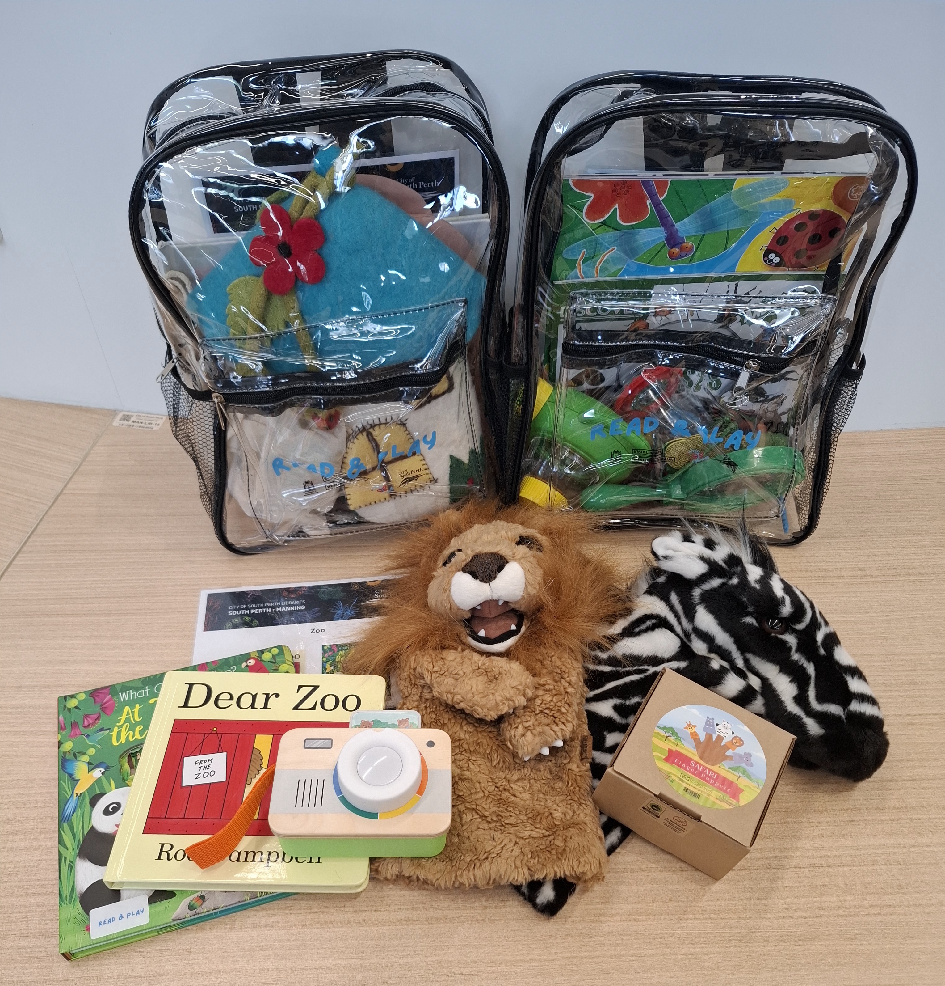Two backpacks filled with books, puppets and headphone with similar contents displayed on a table in front.