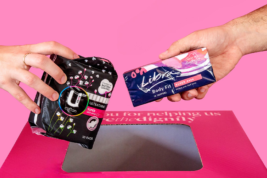 A hand holding a pack of pads and another holding a pack of tampons above a pink donation bin.