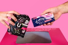 A hand holding a pack of pads and another holding a pack of tampons above a pink donation bin.